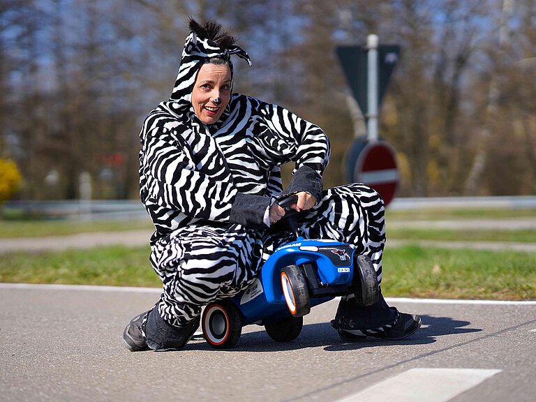 Eine als Zebra verkleidete Frau sitzt auf einem Rutschfahrzeug auf einem Verkehrsübungsplatz.