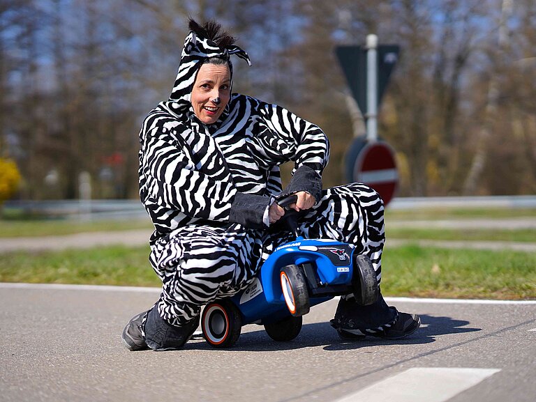 Eine als Zebra verkleidete Frau sitzt auf einem Rutschfahrzeug auf einem Verkehrsübungsplatz.