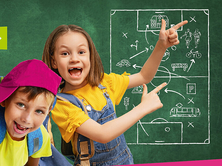 Zwei Schulkinder stehen vor einer Tafel und grinsen in die Kamera.