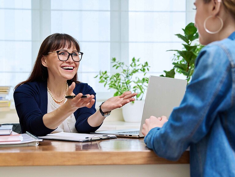 Eine Frau sitzt mit Laptop am Schreibtisch und berät eine andere sitzende Frau.