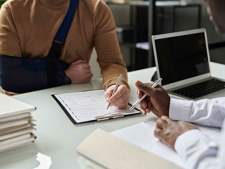 Zwei Personen besprechen etwas an einem Schreibtisch. Eine Person trägt einen Verband am Arm.