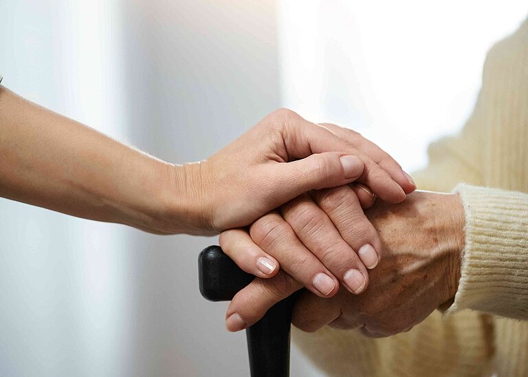 Zwei Händen greifen nacheinander. Es handelt sich um eine jüngere und eine ältere Hand mit Gehstock.