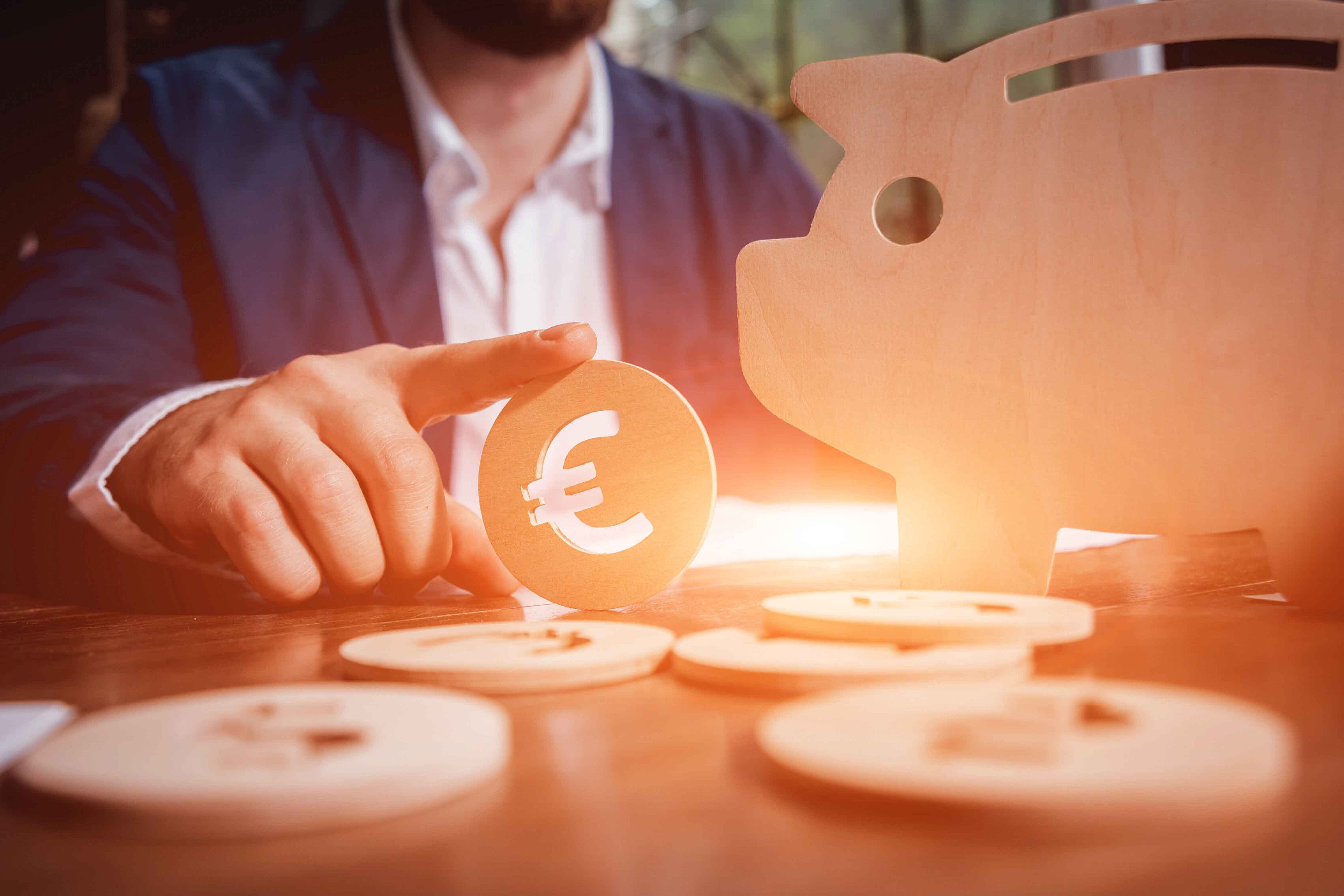 Ein Mann sitzt an einem Tisch. Vor ihm sind Euromünzen aus Holz und ein Schweinchen aus Holz zu sehen.