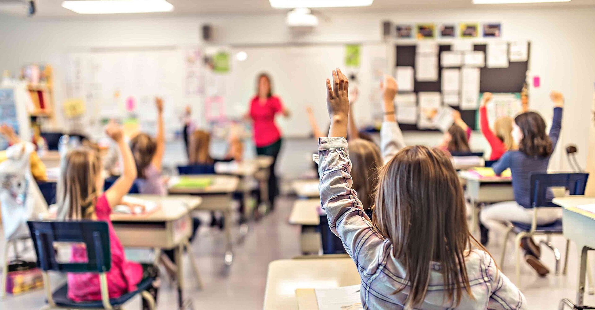 Mehrere Schülerinnen und Schüler befinden sich im Klassenzimmern und haben Unterricht. Einige Schüler melden sich.