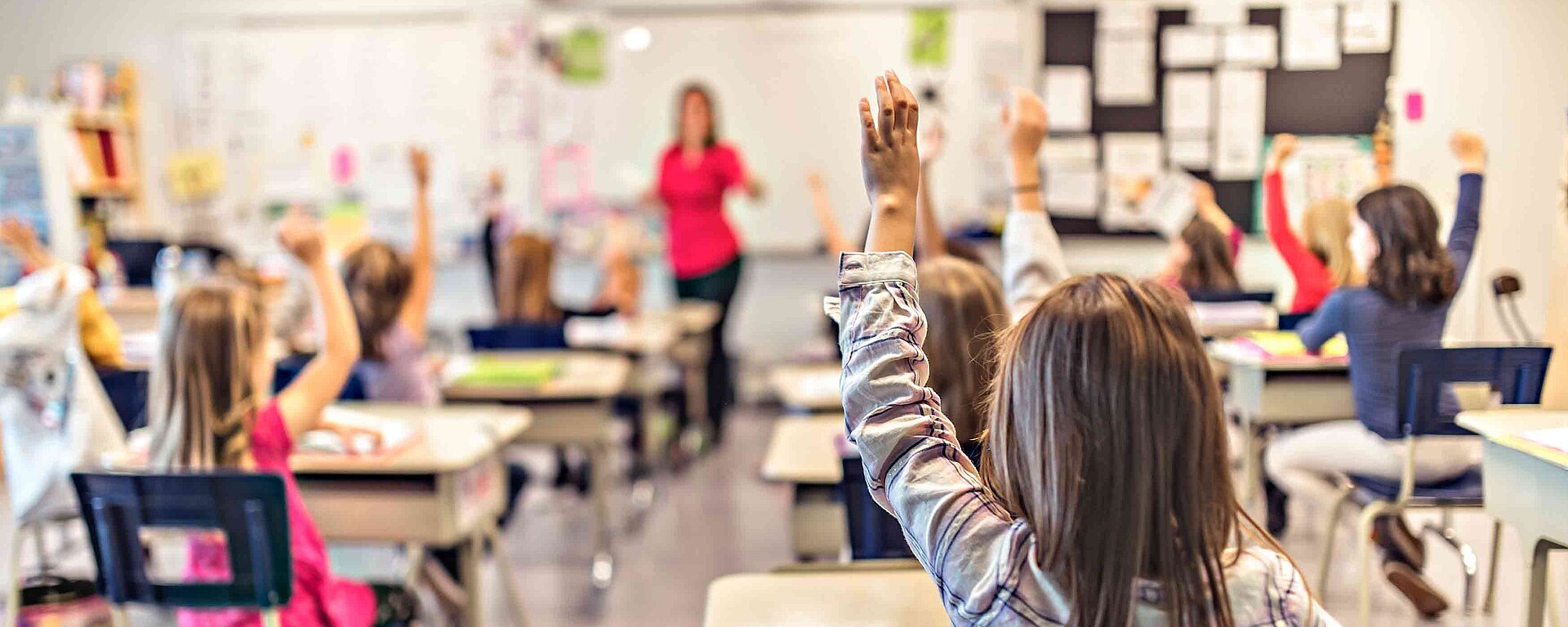 Mehrere Schülerinnen und Schüler befinden sich im Klassenzimmern und haben Unterricht. Einige Schüler melden sich.