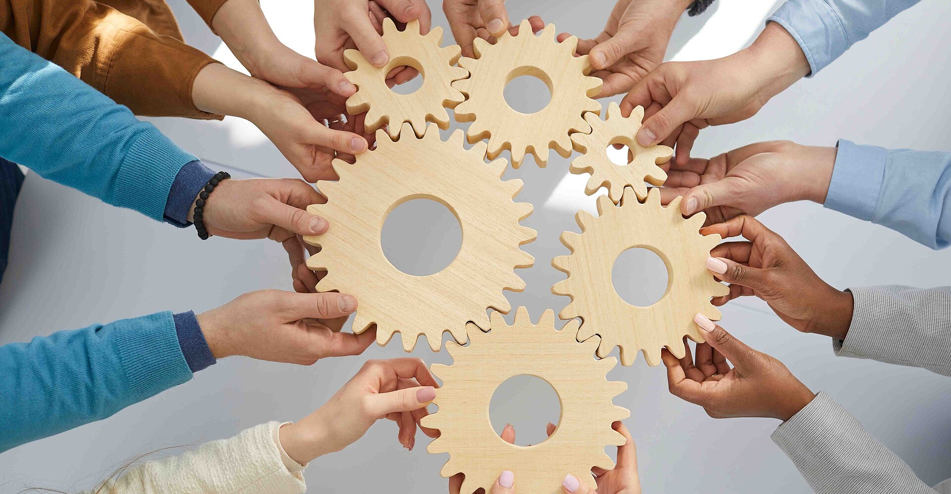 Verschiedene Hände heben Zahnräder aus Holz. Die Zahnräder greifen ineinander und bilden ein großes Ganzes.