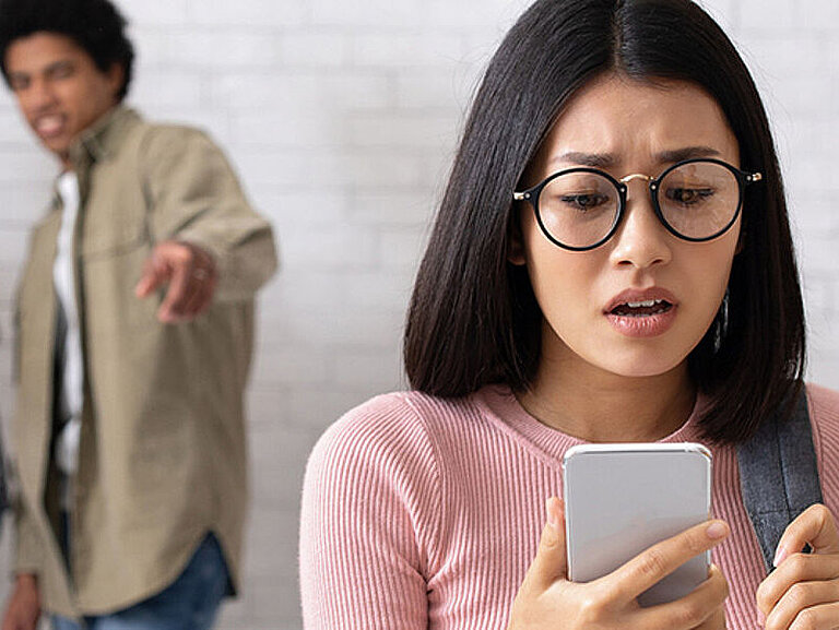 Ein Mädchen schaut verzweifelt auf ihr Smartphone. Im Hintergrund stehen vier Jugendliche und lachen über das Mädchen.