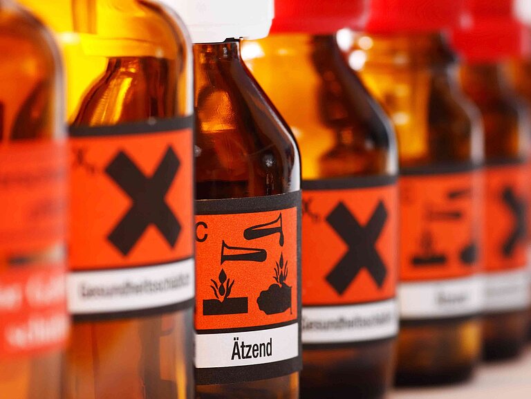 Mehrere Glasflaschen mit orangenen Etiketten. Die Beschilderung zeigt, dass es sich um gefährliche flüssige Stoffe handelt.