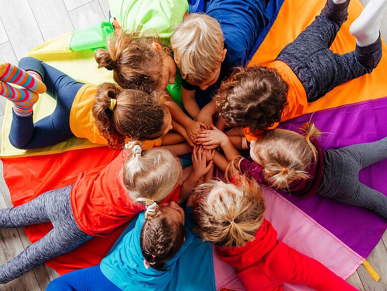 Acht Kinder liegen im Kreis auf einer Decke auf dem Boden und spielen zusammen.