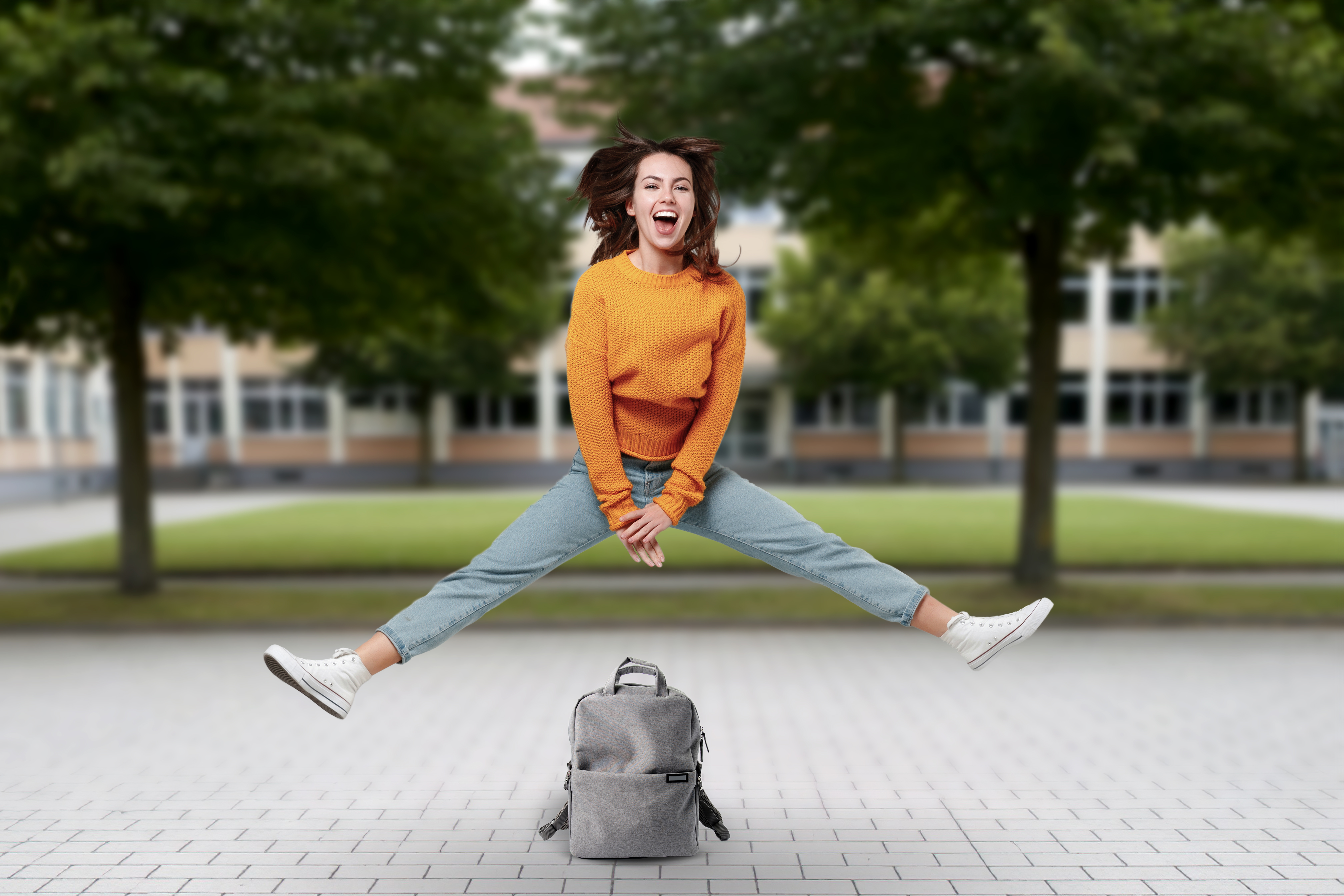 Eine Schülerin springt über ihren Schulrucksack. Sie ist auf einem Pausenhof.